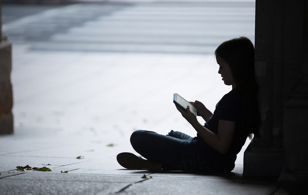 Teen girl sits against wall in shadow, saddened by her iPAd