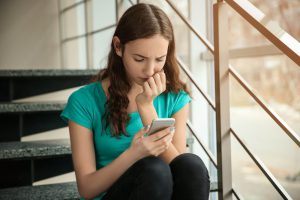 Young girl sits alone on school steps looking at bullying messages on her phone