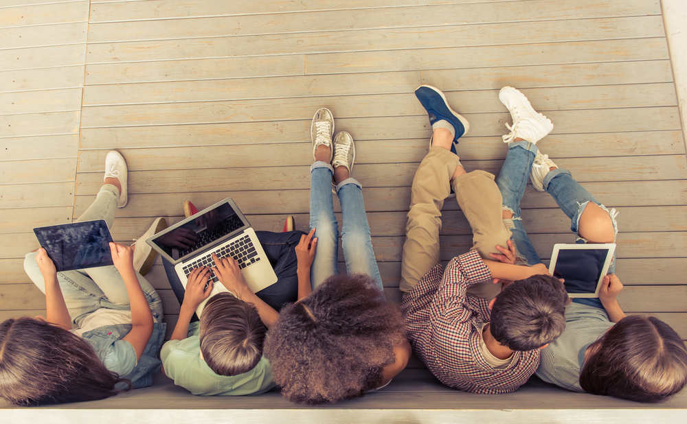 View from above of five teenagers using iPads and laptops while sitting on a wooden floor