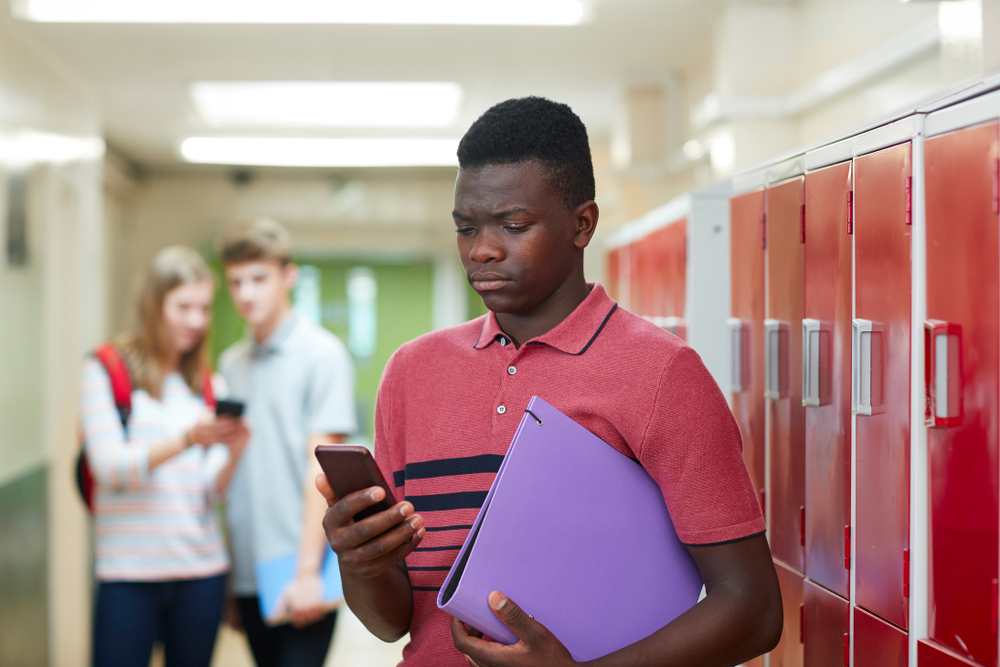Young male holding a binder and looking at his cell phone upset while two other students are laughing in the background in front of some lockers.