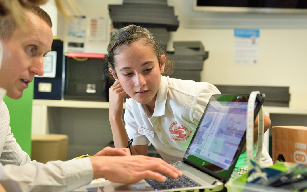 Girl watches teacher use laptop.