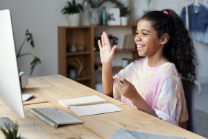 Young girl raises hand at computer while distance learning.