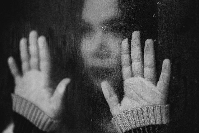 Sad teen girl looks out of window with her hands against the glass.