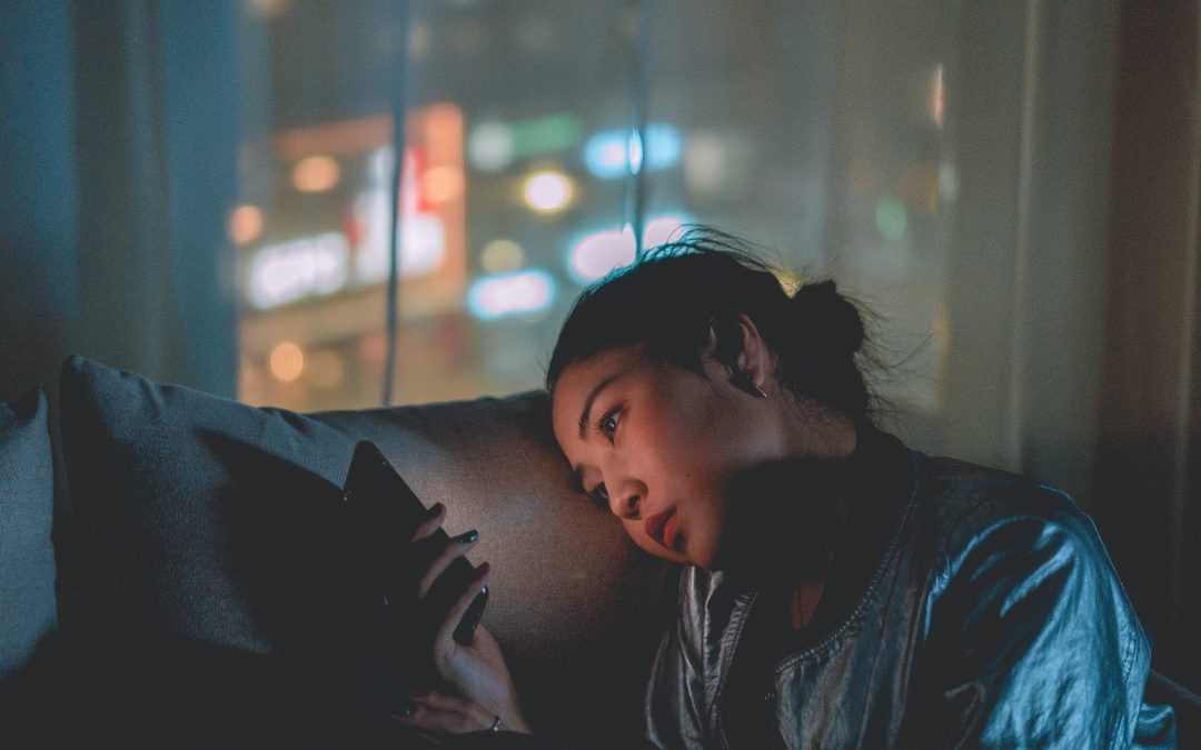 Sad Asian American teen looks at smartphone in a dark room.
