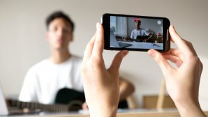 Young man using iPhone to film another young man playing guitar.