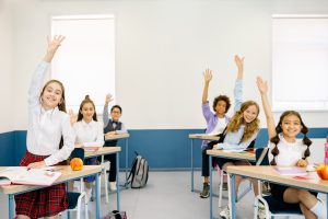 Happy students in desks raising their hands.